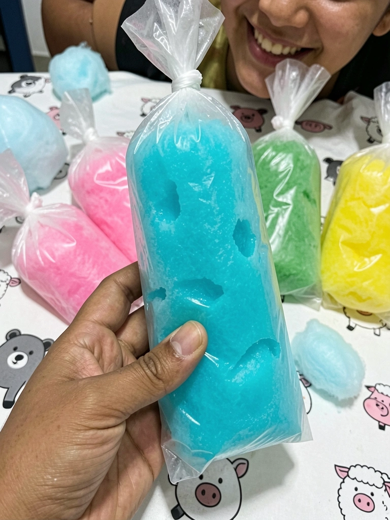 Mão segurando geladinho azul intenso, com outros geladinhos coloridos ao fundo desfocado